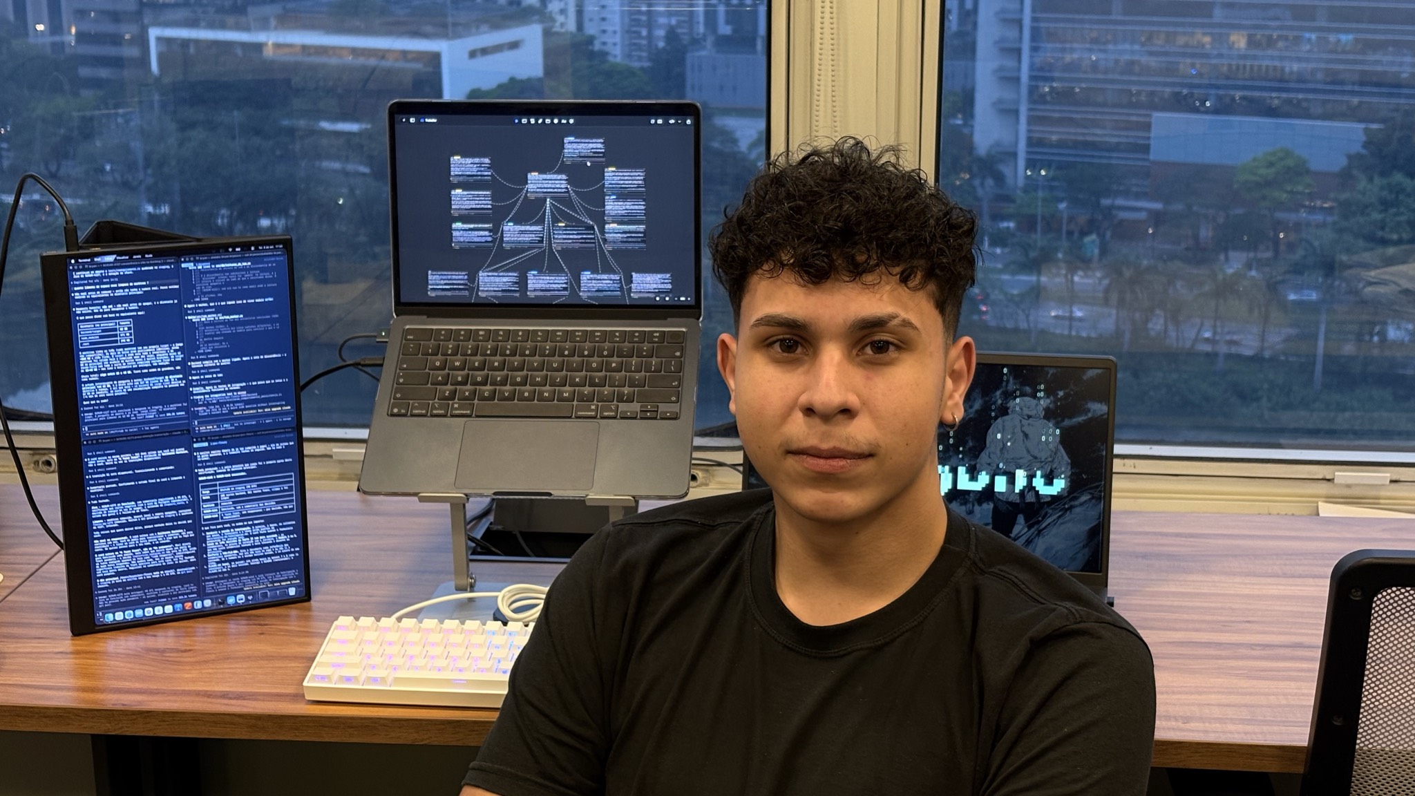 Bryan Pinheiro em sua mesa de trabalho, com computadores e código ao fundo